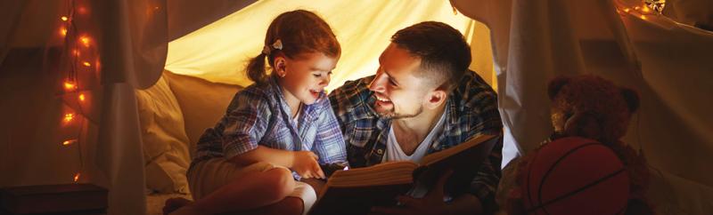 A father and daughter enjoy storytime in a homemade blanket tent with fairy lights.